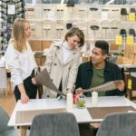 Three young adults discuss table options in a stylish furniture store, symbolizing teamwork and modern interior design.